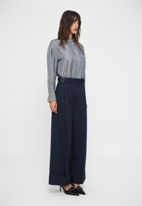 Chemise à rayures bleu et blanc, associée à un pantalon large marine avec une ceinture et des manches repliées, complétée par des chaussures à talons noires.