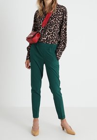 Leopardenmuster Langarmshirt, grüne taillierte Hose, beigefarbene Absatzschuhe und roter Umhängebeutel mit Logo. Glatter Stoff und taillierte Passform.