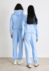 Sudaderas con capucha de color azul claro, acompañadas de pantalones de chándal a juego, que cuentan con puños elásticos y un ajuste relajado. El fondo es neutro y tiene una textura suave.