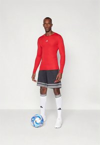 Piros hosszú ujjú sportfelső, szürke rövidnadrág vízszintes csíkokkal, fehér térdig érő zokni fekete részletekkel, fehér futballcipő és színes focilabda.