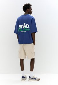 T-shirt oversized in cotone blu con stampa grafica sul retro, abbinato a pantaloni cargo beige. Il modello indossa calze bianche e sneaker grigie.
