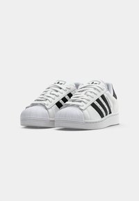 Pár bielych tenisiek Adidas Superstar s čiernymi pruhmi a bielymi šnúrkami, ukázaných z mierneho uhla na bielom pozadí.