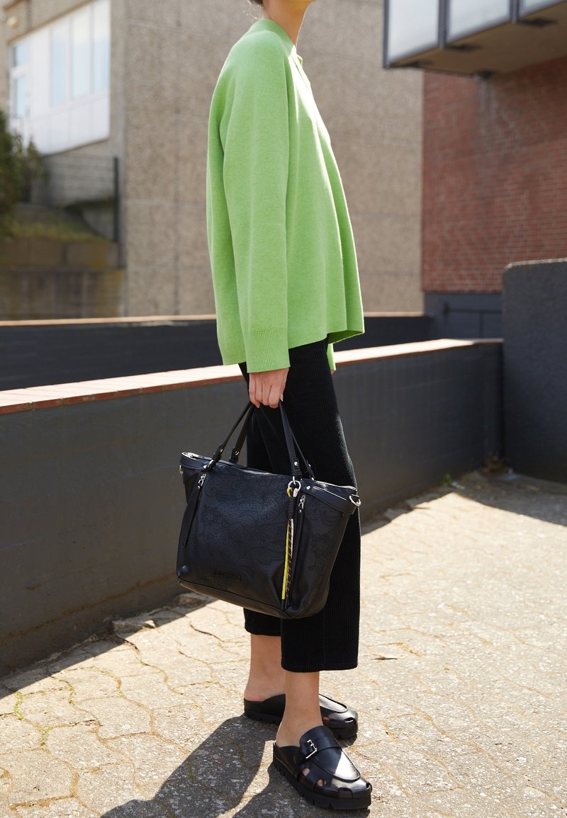 Personne portant un pull vert, un pantalon noir et des sandales noires tient un grand sac fourre-tout noir en se tenant à l'extérieur par une journée ensoleillée.
