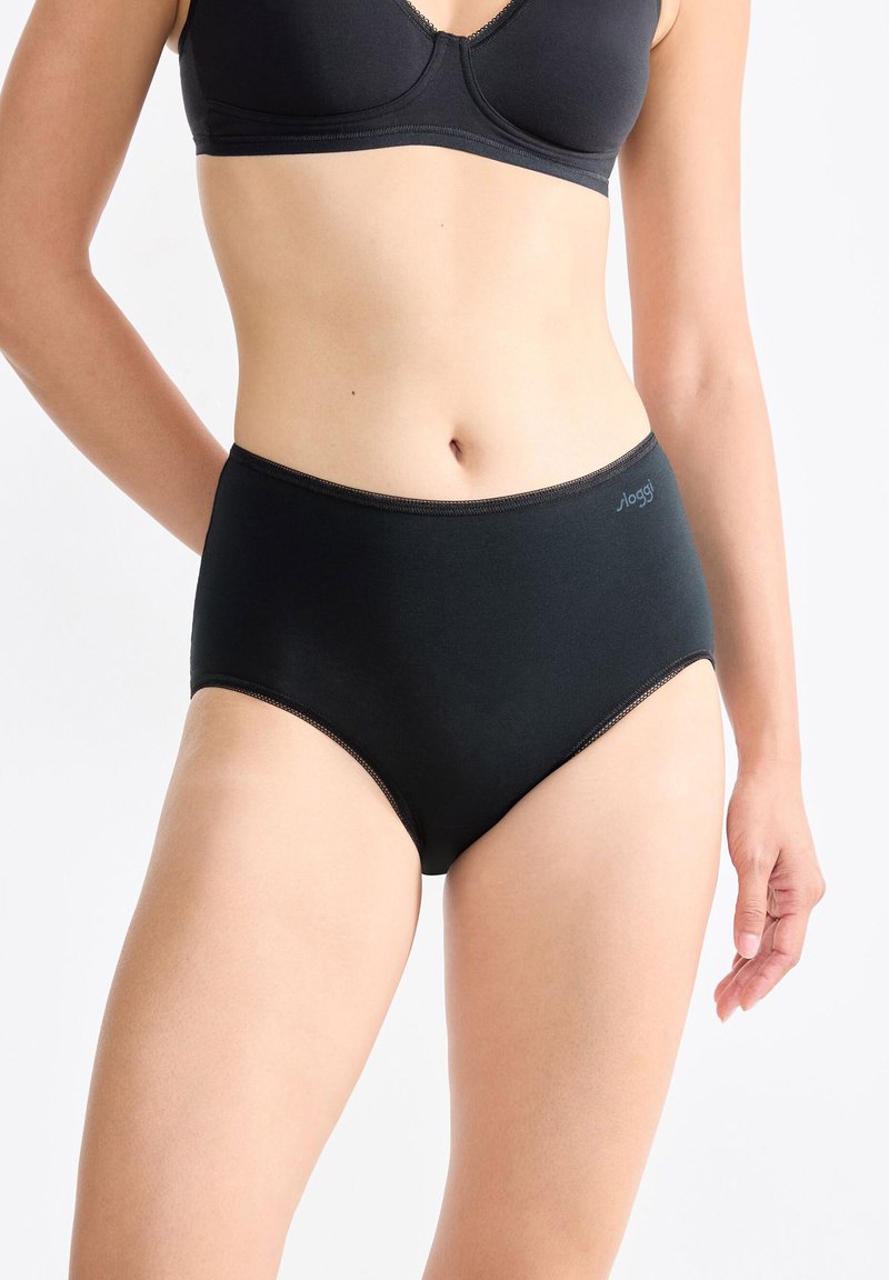 Donna che indossa slip neri a vita alta e un reggiseno nero abbinato su uno sfondo bianco semplice, con le mani rilassate ai fianchi.