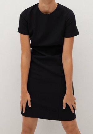 Femme portant une robe noire simple à manches courtes, debout les mains posées près des cuisses sur un fond uni clair.