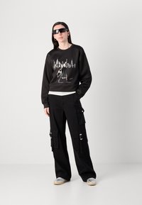 Sweatshirt preto com gráfico de paisagem urbana, combinado com calças cargo pretas que têm vários bolsos e detalhes metálicos. Ténis transparentes completam o visual.