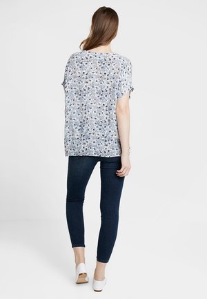 Bluse mit Blumenmuster und kurzen Ärmeln, hellblauer Hintergrund und schwarze Akzente. Kombiniert mit dunkelblauen Skinny-Jeans und weißen Schuhen.