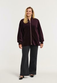 Velvet maroon bomberjakke med lynlås foran, elastiske manchetter og to frontlommer, parret med sorte breddebenede jeans og sorte flade sko.