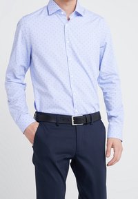 Camisa de botones azul claro con pequeños puntos rojos y azules marinos, de mangas largas y cuello clásico, combinada con pantalones oscuros y un cinturón negro.