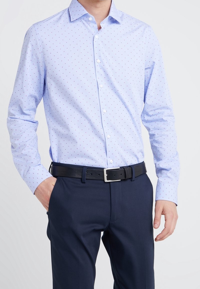 Camisa de botones azul claro con pequeños puntos rojos y azules marinos, de mangas largas y cuello clásico, combinada con pantalones oscuros y un cinturón negro.