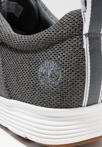 Šedé síťované tenisky s bílou gumovou podrážkou, texturovaným svrškem a modrou vyšitou logem stromu na patě, doplněné bílými a šedými pruhy.