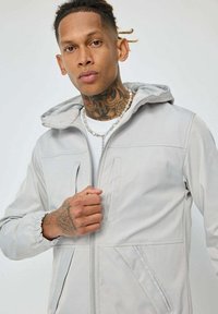 Veste à capuche gris clair avec fermeture éclair, poignets élastiques et deux poches avant. Texture de tissu lisse et design minimaliste.