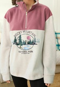 Polarowy pulower w kolorze różowym i kremowym, z półzamkiem. Na przodzie grafika przedstawiająca Park Narodowy Gór Skalistych z drzewami i rzeką.