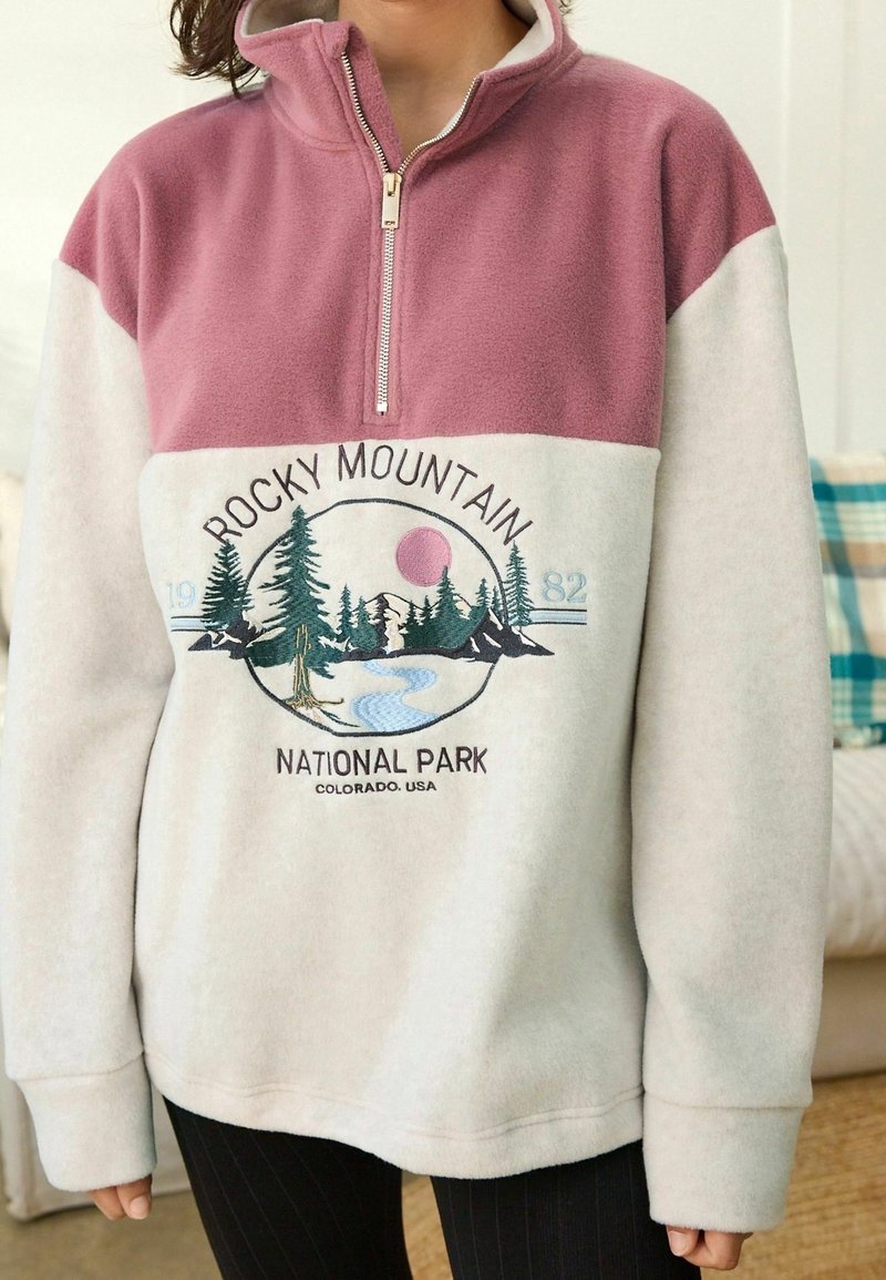 Polarowy pulower w kolorze różowym i kremowym, z półzamkiem. Na przodzie grafika przedstawiająca Park Narodowy Gór Skalistych z drzewami i rzeką.