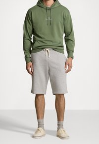 Zelená mikina s kapucí a stahovací šňůrkou, přední kapsa a vyšívaný detail; šedé šortky s bočními kapsami; béžové tenisky a šedé ponožky.