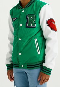 Vihreä ja valkoinen yliopiston takki, jossa on ribbauskainalot, brodeerattu "R"-patchi ja koripallopatchi vasemmassa hihassa. Kaksi sivutaskua.