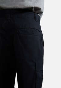 Gros plan d'une personne portant un pantalon cargo bleu marine foncé avec des poches latérales et arrière, associé à une chemise gris clair rentrée.