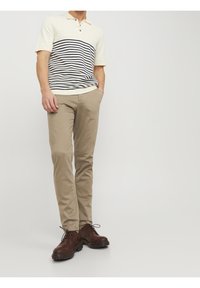 Uomo che indossa un polo crema con strisce navy, pantaloni beige e stivali stringati marrone scuro, in piedi con una mano in tasca.
