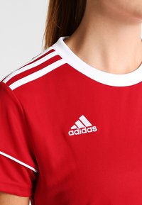 Röd idrottströja med vit krage och tre horisontella vita ränder på ärmarna. Har en broderad Adidas-logotyp på bröstet.