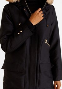 Zwarte parka met een capuchon van namaakbont, ritssluitingen aan de zijkanten en gouden accenten. Heeft een gladde textuur en verstelbare trekkoorden.