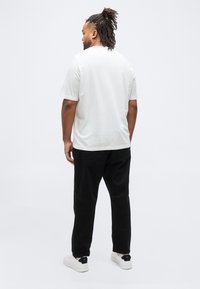 Άνδρας με ράστα στέκεται γυρισμένος πίσω φορώντας απλό λευκό κοντομάνικο πουκάμισο, μαύρο παντελόνι και λευκά αθλητικά παπούτσια σε λευκό φόντο.