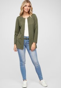 Olive green, textured cardigan with open front, worn over a white top. Paired with light blue jeans and white sneakers.