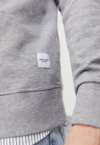 Gray sweatshirt with ribbed cuffs and hem, featuring a small white label with text. Layered over a striped shirt. Soft fabric texture.