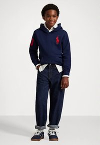 Αγόρι που φοράει ναυτικό φούτερ με κόκκινο λογότυπο, λευκή μπλούζα, σκούρα τζιν με γυρισμένα μπατζάκια και ναυτικά αθλητικά παπούτσια με λευκές κορδόνες, που στέκεται σε απλό φόντο.