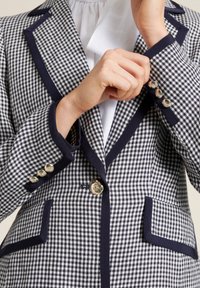 Blazer a cuadros con un patrón en azul marino y blanco, con ribetes en azul marino a contraste, botones dorados y un ajuste entallado con detalles de bolsillos.