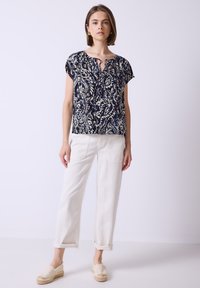 Donna che indossa una blusa corta a maniche corte con motivo blu navy, pantaloni bianchi arrotolati e scarpe espadrillas beige, in piedi contro uno sfondo chiaro uniforme.