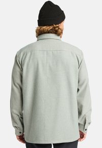 Osoba z kręconymi włosami nosząca czarny, dziany beanie oraz zielono-białą koszulę z długim rękawem w kratkę, widoczna od tyłu.
