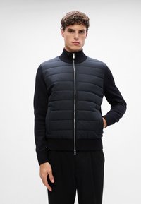 Melnā pūkaino veste ar pāršūtu dizainu, stāvošo apkakli un rāvējslēdzēja aizdari. Piedurknes izgatavotas no gluda, tumša auduma. Valkāta virs melnajām biksēm.