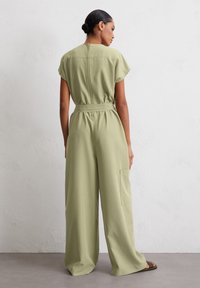Giacca ampie di colore verde chiaro con maniche corte, vita cinchata e tasca laterale. Tessuto liscio con una leggera lucentezza e dettagli con zip sul retro.