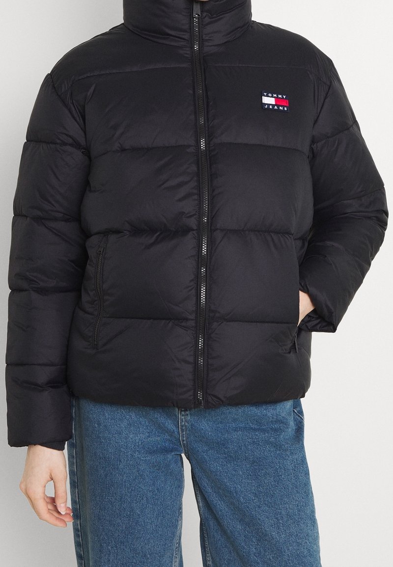 Black puffer jacket with a high collar, front zipper, and padded sections. Features a small logo patch on the left chest.
