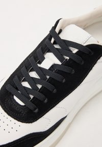 Black and white sneakers with a suede and leather upper, round toe, black laces, perforated toe box, and subtle stitching details.