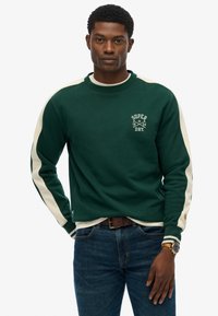 Sudadera verde con rayas blancas en los hombros, que presenta un logo y un tejido texturizado. Combinada con jeans oscuros y un cinturón marrón, el modelo está de pie.