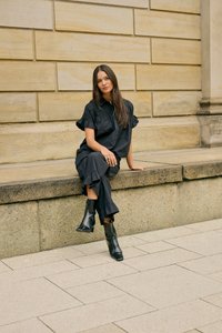Femme aux longs cheveux foncés portant une robe noire et des bottines noires, assise sur un rebord en pierre contre un mur en briques beige.