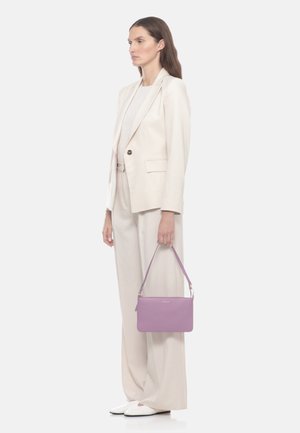 Donna in completo crema e scarpe bianche che tiene una borsa lavanda per la tracolla, in piedi davanti a uno sfondo bianco uniforme.
