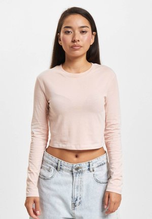 Tânără cu păr lung și închis la culoare, poartă un tricou roz deschis croped cu mâneci lungi și blugi cu talie înaltă, spălați deschis, stând pe un fundal alb.