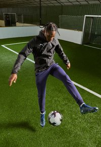 Atletická postava v čiernej bunde a fialových nohaviciach, kopajúca do vzorovanej futbalovej lopty na umelom trávniku v vnútornom zariadení.