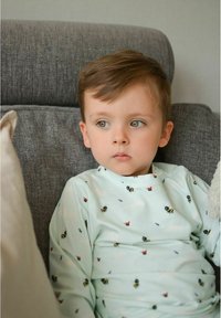 Ljusblå långärmad tröja med små mönster av bin och fjärilar. Barnet sitter på en grå soffa med kuddar i neutrala färger.