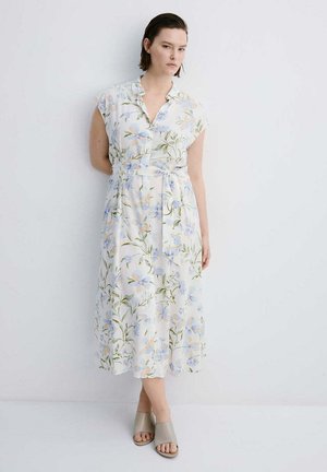 Mujer con cabello corto y castaño, vistiendo un vestido midi blanco con flores y mangas tipo capa, y zapatos abiertos beige sin cordones, de pie frente a una pared blanca.