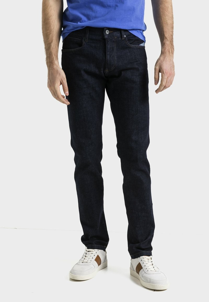 Pantalones vaqueros de denim azul oscuro con un corte ajustado, cinco bolsillos y cierre de botón. Combinados con zapatillas de color claro y una camisa azul.