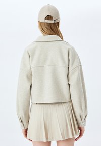 Kurz geschnittene, hellgraue Jacke mit Kragen und geknöpften Manschetten, kombiniert mit einem plissierten, beigen Rock. Eine graue Mütze rundet den Look ab.