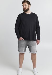 Chemise noire à manches longues avec un col rond, associée à un short en denim gris. Le modèle porte des baskets blanches. Design simple et épuré.