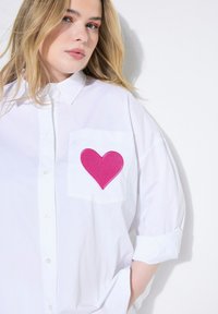 Hvid oversized skjorte med en pink hjerte-applikation på venstre lomme. Har en klassisk krave og knaplukning foran, lavet af glat stof.