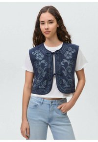 Temno moder izvezen jopič z cvetličnimi vzorci, okroglim ovratnikom in sprednjim zapenjanjem, oblečen čez belo srajco in svetlo kavbojke.