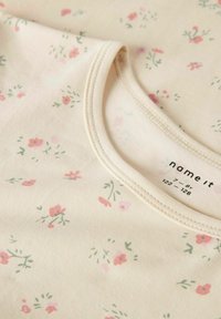 Hellbeige Baumwollhemd mit floralem Druck in Pink und Grün. Ausgerundeter Ausschnitt mit sichtbaren Nähten und Markenlabel innen.