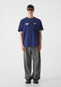 Mörkblå bomullströja med rosa text på bröstet, kombinerad med lösa grå byxor och svarta platåskor. Enkel bakgrund.