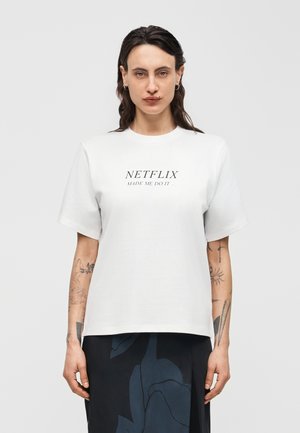 Personne aux longs cheveux foncés portant un t-shirt blanc avec l'inscription « NETFLIX MADE ME DO IT » et une jupe à motifs sombres, debout devant un fond uni.